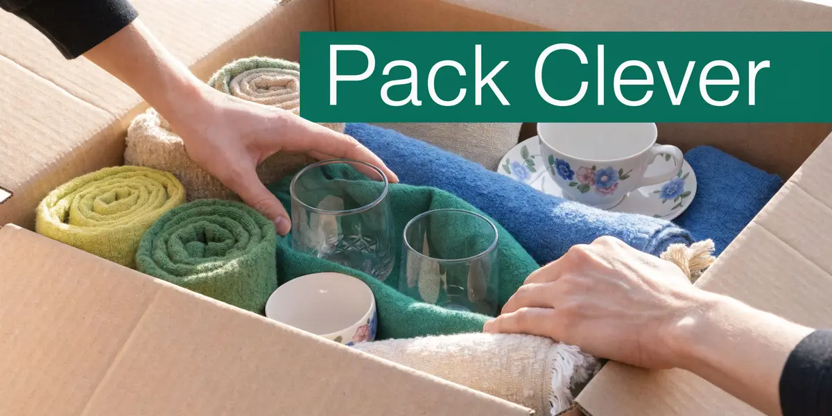 A person carefully packing household items like glasses and dishes into a cardboard moving box using towels.